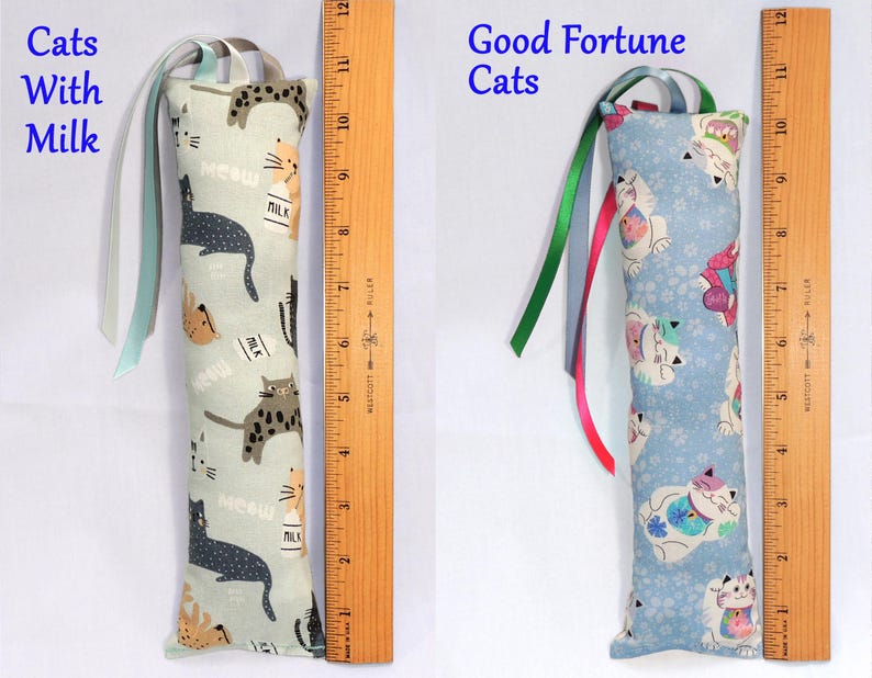 May include: Two cat-themed catnip toys with ribbons. The first toy is light blue with a pattern of black cats and milk cartons. The second toy is light blue with a pattern of white cats with good fortune symbols. Both toys are approximately 10 inches long.