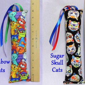 May include: Two cat-themed catnip toys. The first toy is a long, rectangular pillow with a colorful print of cats with rainbow colors. The second toy is a long, rectangular pillow with a black background and a print of sugar skull cats. Both toys have long ribbons attached.
