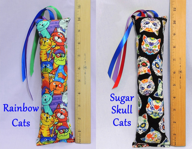 May include: Two cat-themed catnip toys. The first toy is a long, rectangular pillow with a colorful print of cats with rainbow colors. The second toy is a long, rectangular pillow with a black background and a print of sugar skull cats. Both toys have long ribbons attached.