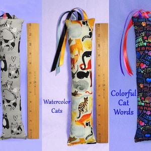 May include: Three cat toys with colourful ribbon accents. The first is grey with black and white cat designs. The second has watercolour cat designs. The third is black with colourful cat-themed words. Each toy is approximately 30 cm long.