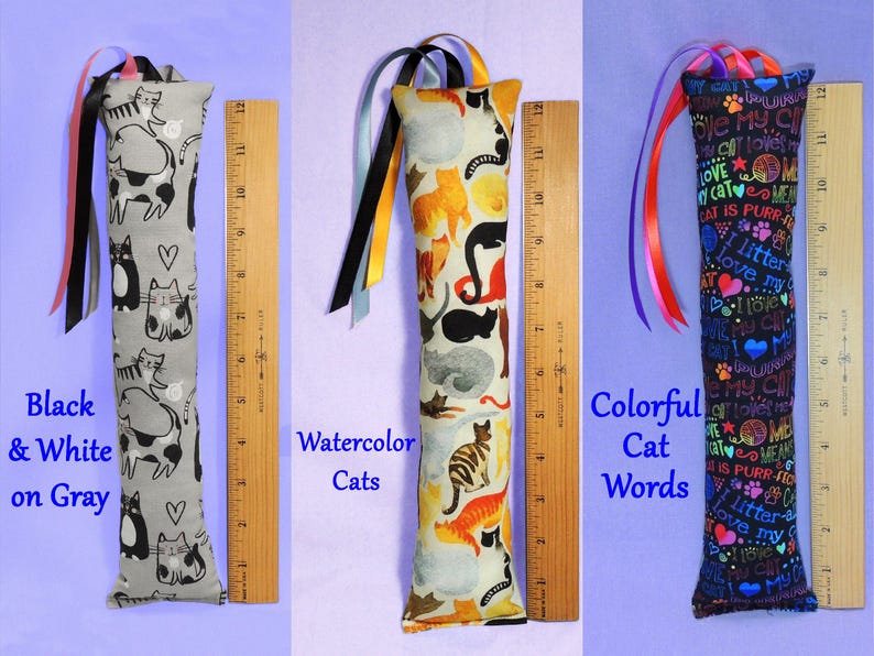 May include: Three cat toys with colourful ribbon accents. The first is grey with black and white cat designs. The second has watercolour cat designs. The third is black with colourful cat-themed words. Each toy is approximately 30 cm long.