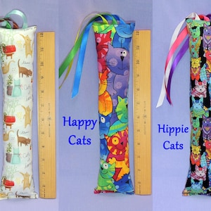 May include: Three cat toy tubes, each approximately 30 cm long, with vibrant cat-themed fabric patterns. One reads "Cats at Home," another "Happy Cats," and the third "Hippie Cats." They all feature ribbon embellishments.