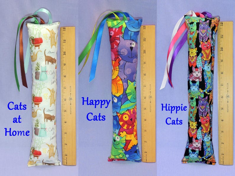 May include: Three cat toy tubes, each approximately 30 cm long, with vibrant cat-themed fabric patterns. One reads "Cats at Home," another "Happy Cats," and the third "Hippie Cats." They all feature ribbon embellishments.