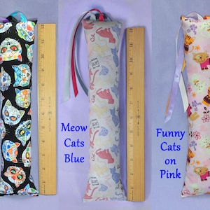 May include: Three cat toys with patterned fabric. The first features sugar skull cat faces on black, the second has blue and pink cat illustrations on white, and the third has orange cat illustrations on pink. Each toy is about 30 cm long and has a ribbon attached.