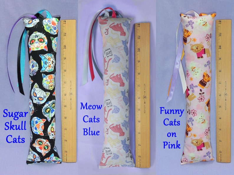 May include: Three cat toys with patterned fabric. The first features sugar skull cat faces on black, the second has blue and pink cat illustrations on white, and the third has orange cat illustrations on pink. Each toy is about 30 cm long and has a ribbon attached.