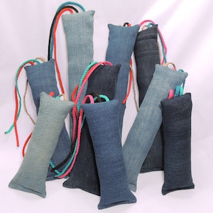 May include: A collection of rectangular denim fabric pouches in various shades of blue, ranging from light to dark indigo. Each pouch features a braided cord loop in bright colors including red, pink, black, and turquoise attached to the top edge.