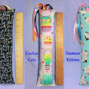 May include: Three cat toys with colourful ribbon accents. One is black with white cat faces, one is pink with cactus cat designs, and the last is light blue with Siamese kitten patterns. Each toy is approximately 30 cm long.