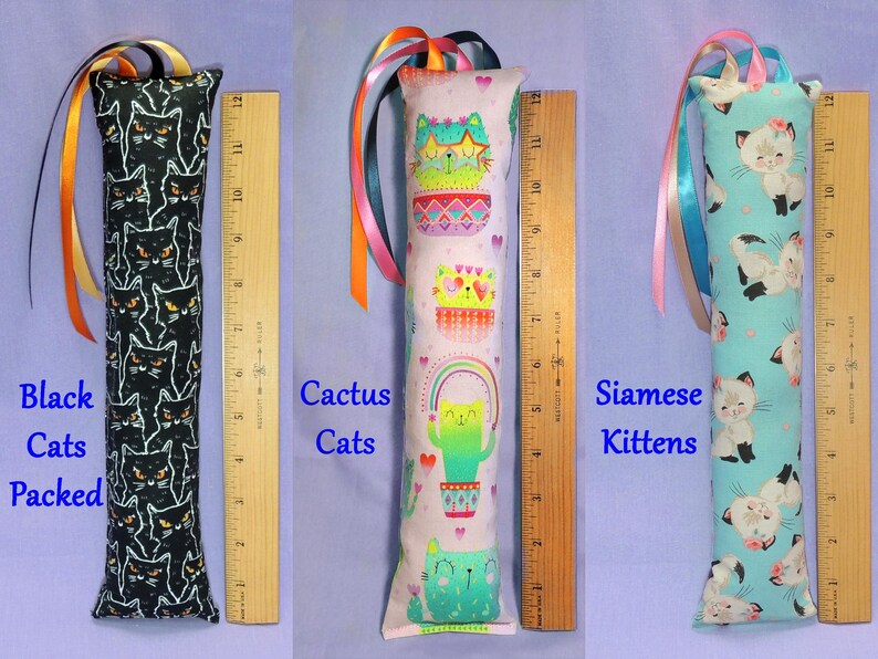 May include: Three cat toys with colourful ribbon accents. One is black with white cat faces, one is pink with cactus cat designs, and the last is light blue with Siamese kitten patterns. Each toy is approximately 30 cm long.