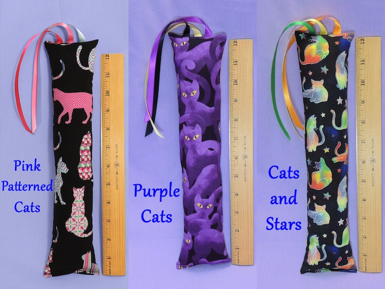 May include: Three cat toy wands with colourful ribbon streamers, each about 30 cm long. One has pink cat patterns on black fabric, another has purple cat designs, and the third has cats and stars on black fabric.