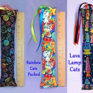 May include: Three cat toys are displayed. One is black with colourful cat doodles, another is packed with rainbow cats, and the last has a lava lamp cat design. Each toy is approximately 30 cm long and has colourful ribbons.