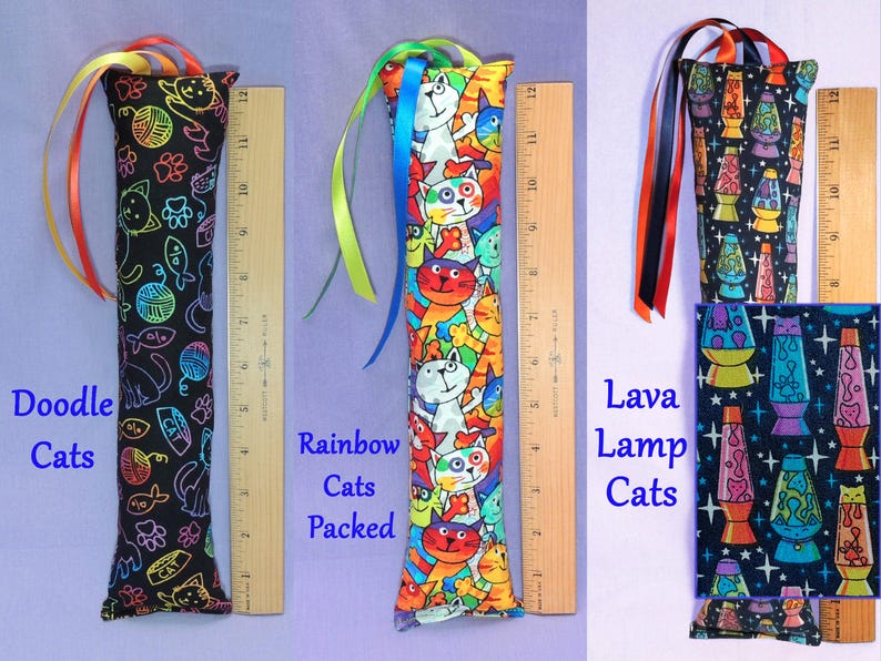 May include: Three cat toys are displayed. One is black with colourful cat doodles, another is packed with rainbow cats, and the last has a lava lamp cat design. Each toy is approximately 30 cm long and has colourful ribbons.