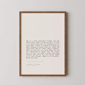 May include: A framed print featuring a quote by Anthony Bourdain in black text on a white background. The quote encourages new experiences. The frame is a dark brown colour.