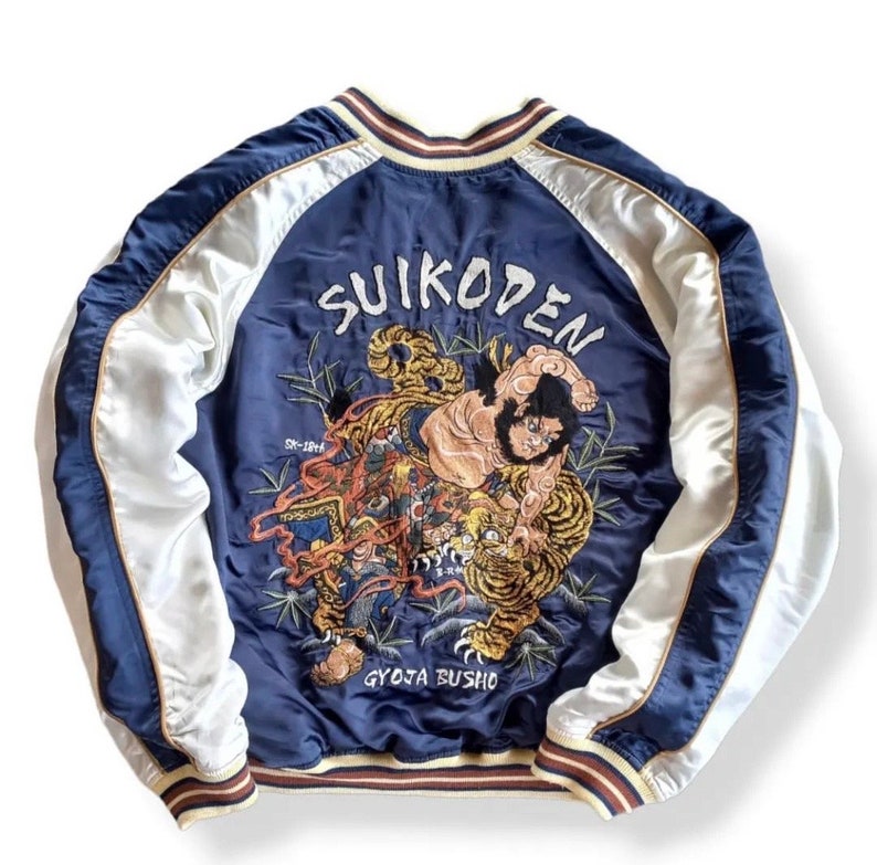 Puede incluir: Una chaqueta bomber de sat&eacute;n azul marino con mangas de sat&eacute;n blanco. La parte posterior de la chaqueta presenta un gran dise&ntilde;o bordado de un tigre y un hombre, con el texto "SUIKODEN" y "GYOJA BUSHO" en caracteres japoneses.