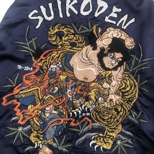 Puede incluir: Una chaqueta bomber azul marino con una imagen bordada de un hombre luchando contra un tigre. La imagen est&aacute; rodeada de bamb&uacute; y el texto "SUIKODEN" est&aacute; bordado en la parte superior. El texto "GYOJA BUSHO" est&aacute; bordado en la parte inferior.