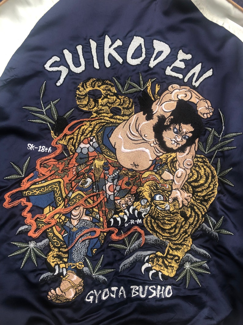 Puede incluir: Una chaqueta bomber azul marino con una imagen bordada de un hombre luchando contra un tigre. La imagen est&aacute; rodeada de bamb&uacute; y el texto "SUIKODEN" est&aacute; bordado en la parte superior. El texto "GYOJA BUSHO" est&aacute; bordado en la parte inferior.