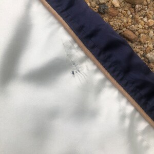 Puede incluir: Una chaqueta blanca con ribete azul marino y beige. La chaqueta tiene un peque&ntilde;o agujero cerca de la costura.