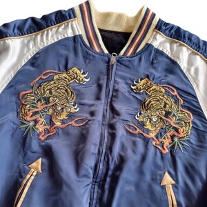 Puede incluir: Una chaqueta bomber azul marino con ribetes de canal&eacute; a rayas blancas y beige alrededor del cuello y los pu&ntilde;os. La chaqueta tiene una cremallera y dos tigres bordados en la parte delantera.