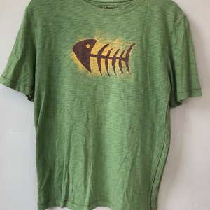 Può includere: T-shirt verde a maniche corte con grafica a lisca di pesce. La lisca di pesce è marrone e gialla. La maglietta è appesa a un appendiabiti di legno. Il marchio "L.L. Bean" è visibile sulla maglietta.