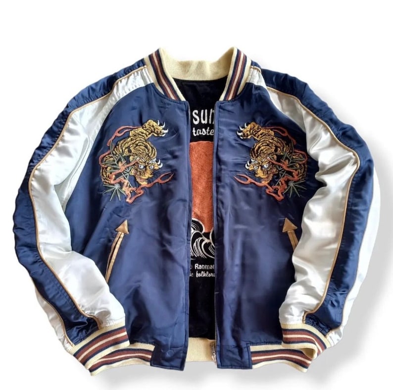 Puede incluir: Una chaqueta bomber de sat&eacute;n azul marino con mangas de sat&eacute;n blanco. La chaqueta presenta tigres bordados en la parte delantera y el texto "Sun" y "Taste" en el lado izquierdo. La chaqueta tiene un cuello y pu&ntilde;os a rayas blanco y marr&oacute;n.