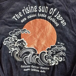 Puede incluir: Una chaqueta negra con un dise&ntilde;o bordado blanco de un sol naciente y olas. El texto "The rising sun of Japan neo asian taste clothing Bakuretsu Rahman Musume eccentric folklore studio SK-19th" est&aacute; bordado en la chaqueta.