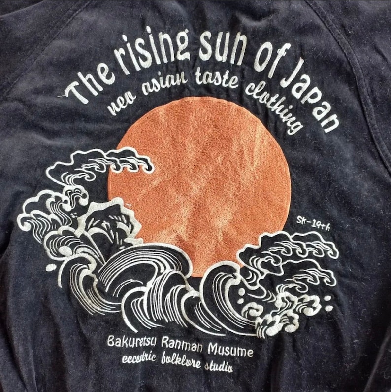 Puede incluir: Una chaqueta negra con un dise&ntilde;o bordado blanco de un sol naciente y olas. El texto "The rising sun of Japan neo asian taste clothing Bakuretsu Rahman Musume eccentric folklore studio SK-19th" est&aacute; bordado en la chaqueta.