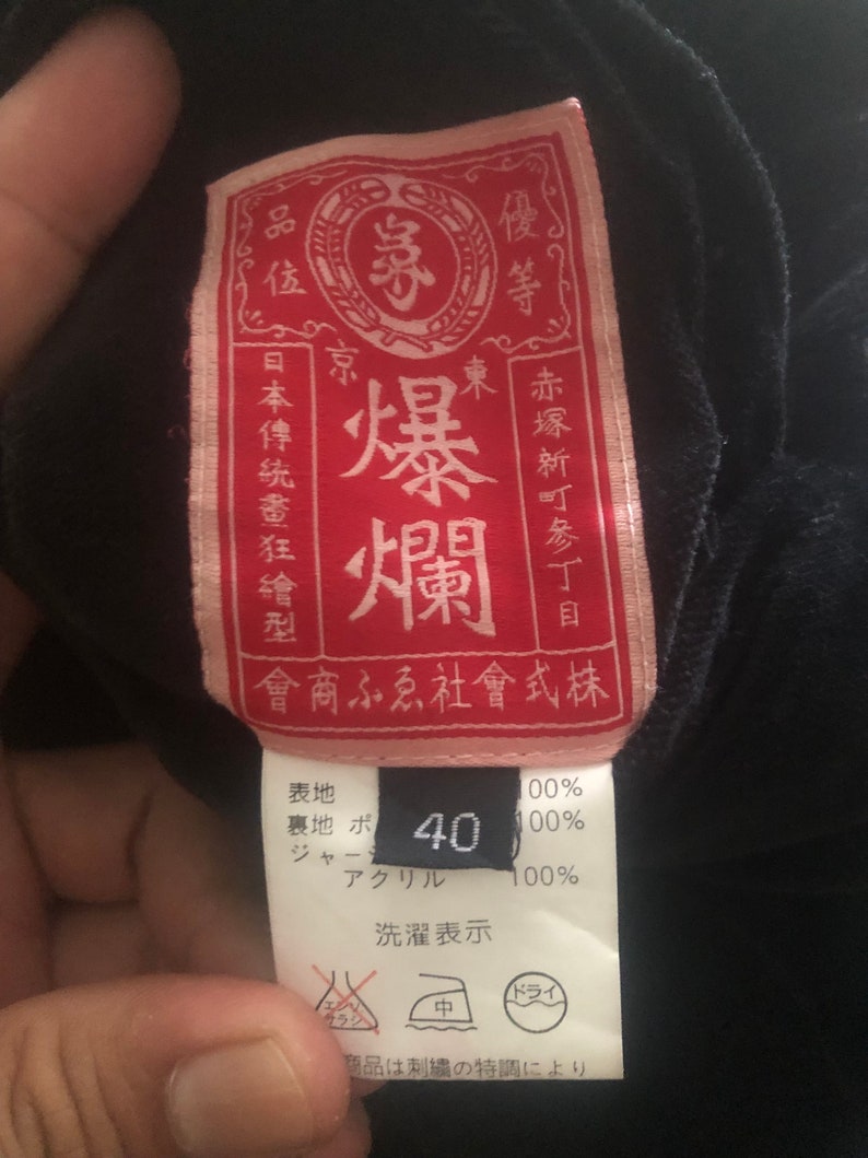 Puede incluir: Una etiqueta de ropa roja y rosa con texto japon&eacute;s que dice "Explosi&oacute;n del Este" y "Quality Goods Co., Ltd.". La etiqueta tambi&eacute;n incluye una etiqueta de talla que dice "40".