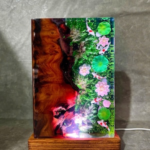 May include: A rectangular resin lamp with a wooden base. The lamp features a koi pond design with white and red fish, pink flowers, and green lily pads. The left side has a wood grain pattern. The lamp is lit from the base.