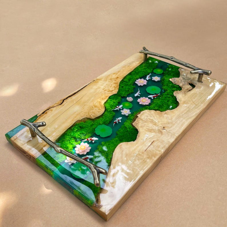 May include: A rectangular wooden serving tray with a river design. The river is made of green resin with white koi fish, pink lotus flowers, and green lily pads. The tray has metal branch handles.