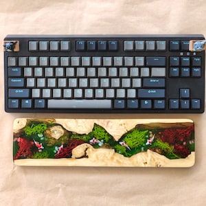 May include: A black mechanical keyboard with a light blue backlight and a wooden keyboard rest with a resin inlay of a stream with green moss and red coral. The resin inlay features white fish.