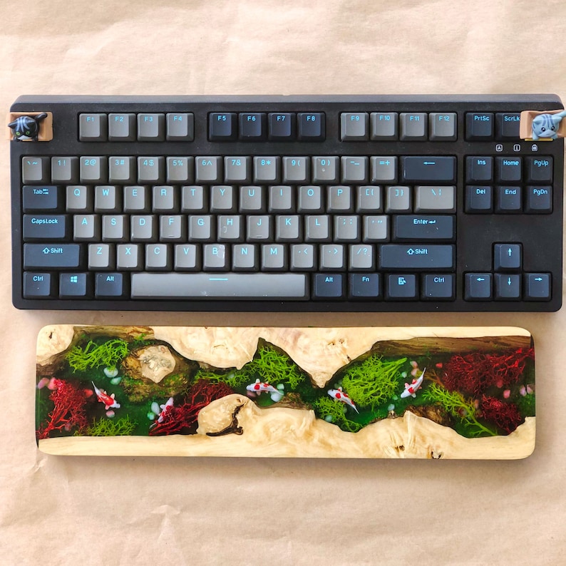 May include: A black mechanical keyboard with a light blue backlight and a wooden keyboard rest with a resin inlay of a stream with green moss and red coral. The resin inlay features white fish.