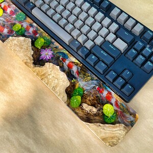 May include: A custom mechanical keyboard with a wooden wrist rest featuring a koi pond design. The wrist rest has a clear resin finish with painted koi fish, lily pads, and a lotus flower. The keyboard has gray and dark blue keycaps.