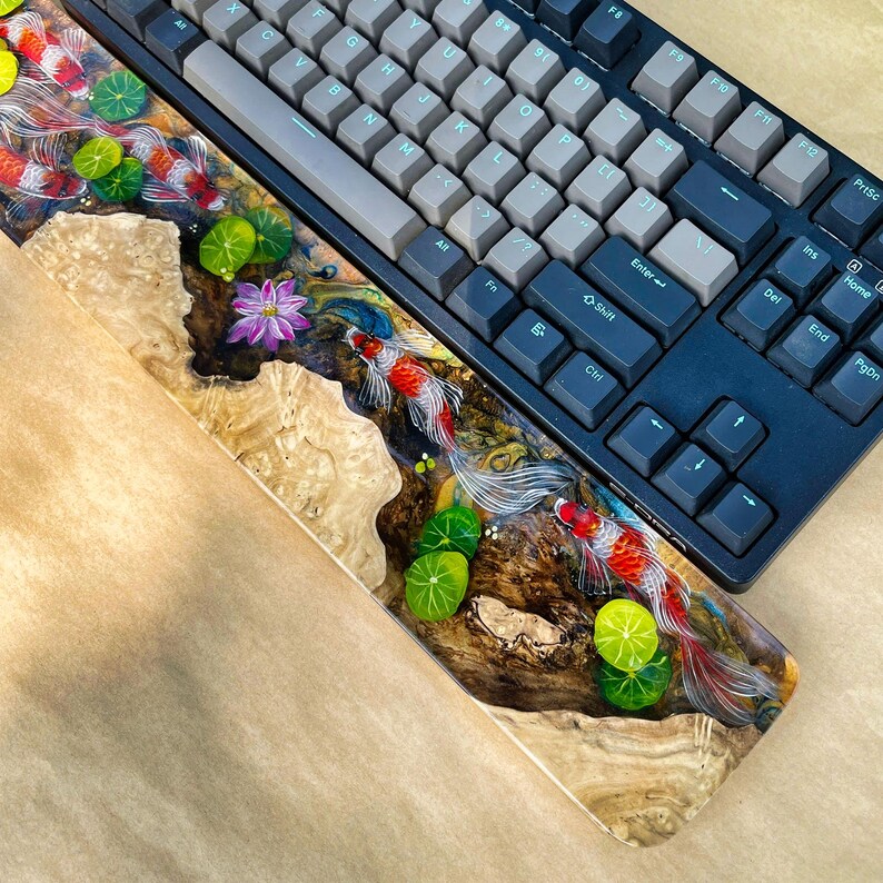 May include: A custom mechanical keyboard with a wooden wrist rest featuring a koi pond design. The wrist rest has a clear resin finish with painted koi fish, lily pads, and a lotus flower. The keyboard has gray and dark blue keycaps.