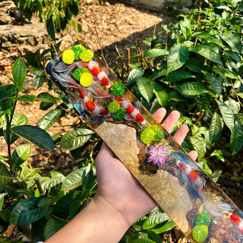 May include: A rectangular wooden art piece featuring a koi pond design. The artwork showcases koi fish with red and white markings swimming among lily pads. The piece is held in a hand, with a natural wood grain base.