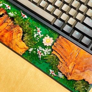 May include: A custom keyboard with a wooden wrist rest featuring a koi pond design. The wrist rest has a green epoxy river with white and red koi fish, white stones, and a pink flower. The keyboard has beige keycaps.