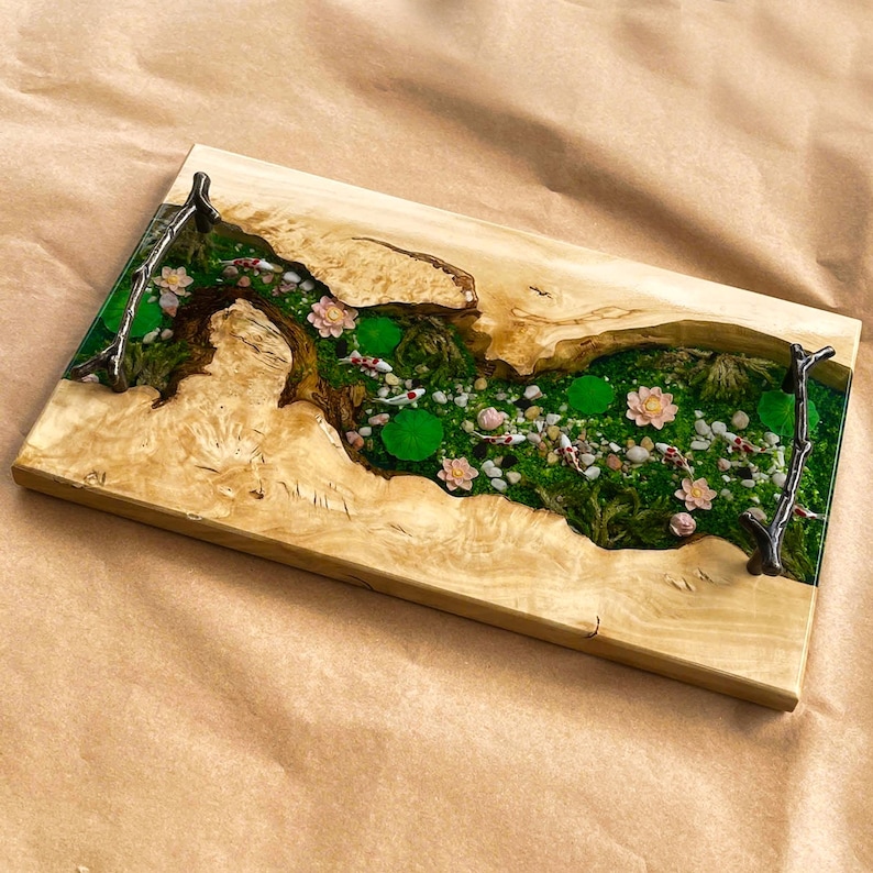 Op de afbeelding: Rechthoekig houten serveerblad met een rivierontwerp. De rivier is gevuld met groen mos, kleine stenen en roze bloemen, met koi-karper. Het blad heeft metalen handgrepen. Afmetingen: ongeveer 40 cm x 20 cm.