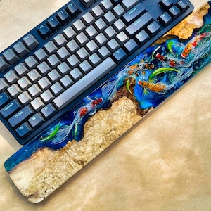 May include: A custom mechanical keyboard with a matching wrist rest. The wrist rest features a vibrant design of koi fish swimming in a blue resin river, set against a wooden base. The keyboard has dark blue keycaps.