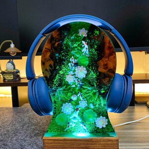 May include: A blue headphone stand with a wooden base. The stand features a resin design with a pond scene, including koi fish, lily pads, and flowers. The headphones are blue.