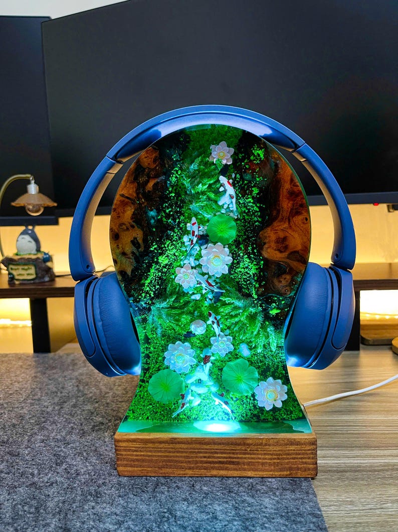 May include: A blue headphone stand with a wooden base. The stand features a resin design with a pond scene, including koi fish, lily pads, and flowers. The headphones are blue.