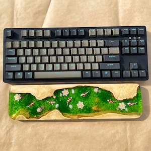 Puede incluir: Un teclado mec&aacute;nico negro con teclas grises se encuentra encima de un reposamu&ntilde;ecas de madera. El reposamu&ntilde;ecas presenta un dise&ntilde;o de resina epoxi verde con flores blancas y peces koi rojos y blancos.