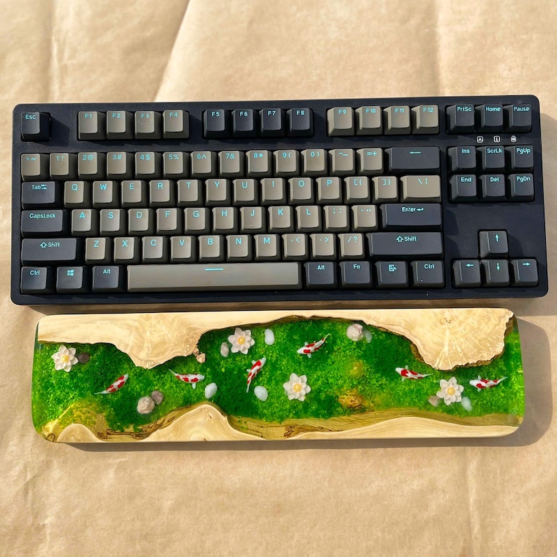 Puede incluir: Un teclado mec&aacute;nico negro con teclas grises se encuentra encima de un reposamu&ntilde;ecas de madera. El reposamu&ntilde;ecas presenta un dise&ntilde;o de resina epoxi verde con flores blancas y peces koi rojos y blancos.