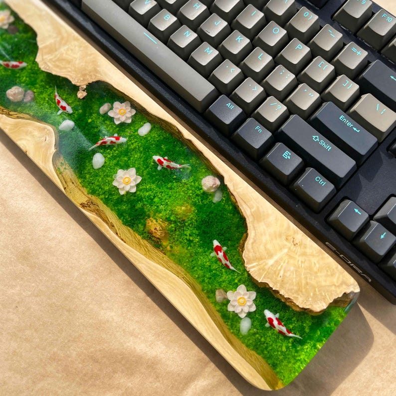 Puede incluir: Un reposamu&ntilde;ecas de teclado de madera con un dise&ntilde;o de r&iacute;o de resina epoxi verde. El r&iacute;o presenta peces koi en miniatura, flores blancas y piedras. Un teclado mec&aacute;nico se encuentra encima.