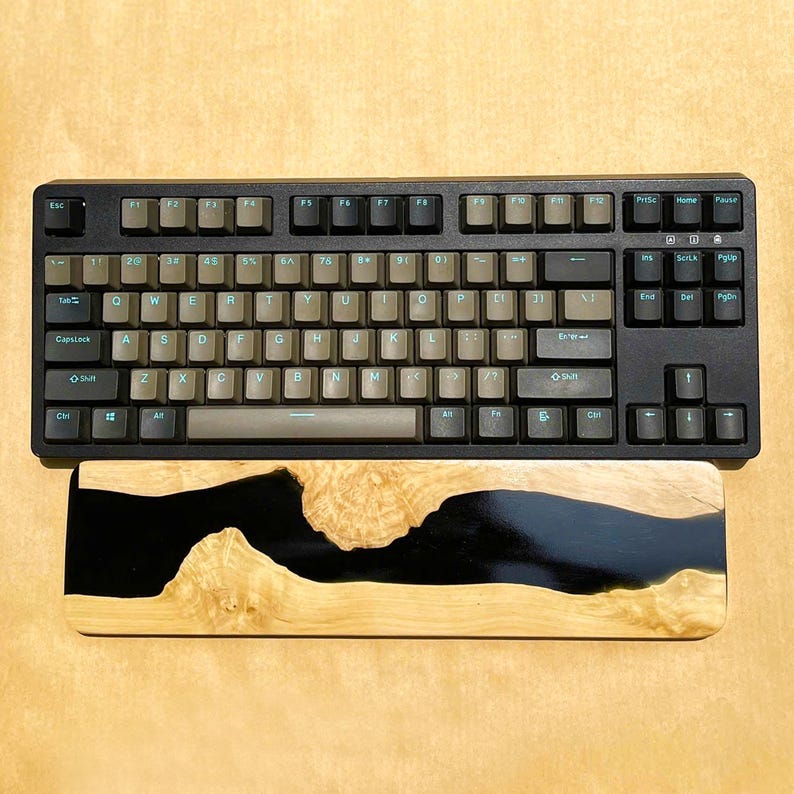 K&ouml;nnte beinhalten: Schwarze mechanische Tastatur mit hellbraunen Tastenkappen und einer h&ouml;lzernen Handgelenkauflage. Die Handgelenkauflage hat ein schwarzes Epoxidharz-Flussdesign mit hellbrauner Holzmaserung. Die Tastatur hat Funktionstasten und einen Ziffernblock.