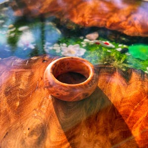 May include: A polished wooden ring with a smooth, rounded band featuring a natural swirling grain pattern in warm shades of amber, orange, and brown, showcasing the organic texture and intricate knots of the wood material.