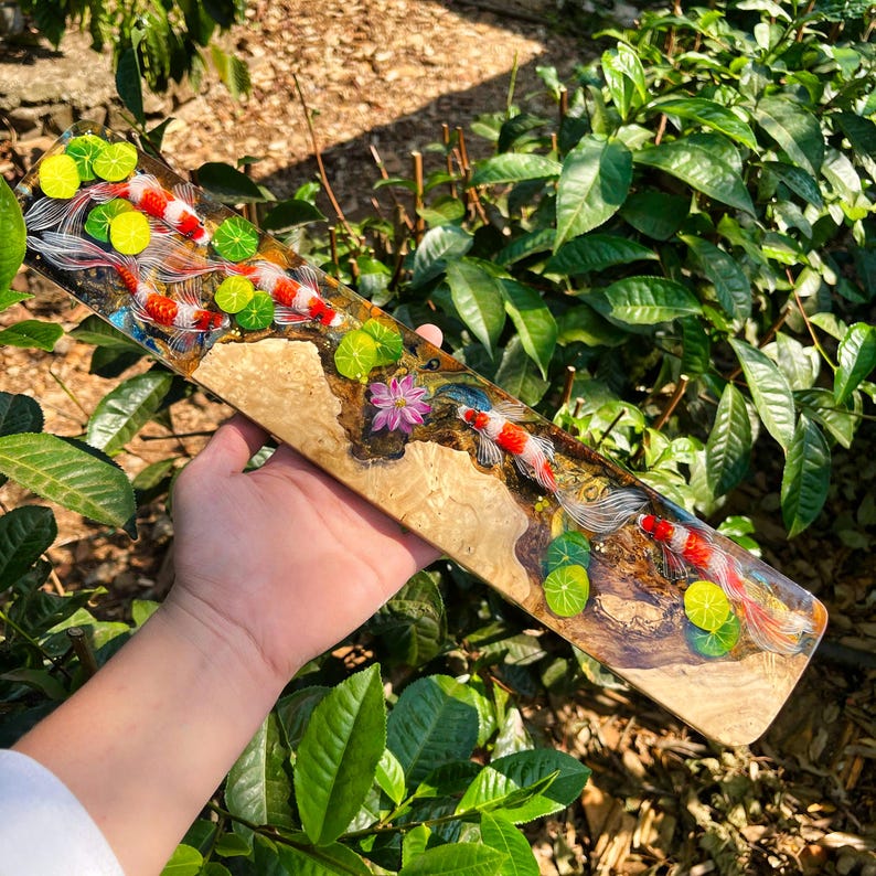 May include: A rectangular wooden art piece featuring a koi pond design. The artwork showcases red and white koi fish swimming among lily pads and flowers. The wood has a natural, light brown grain.