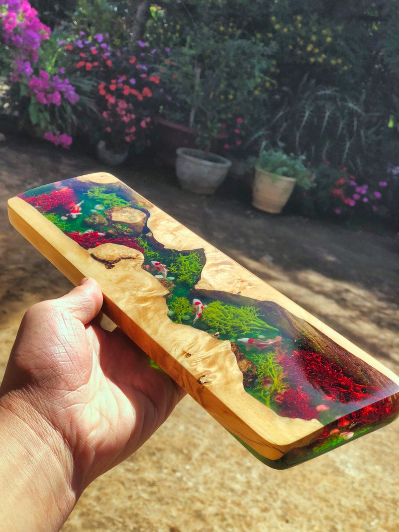 May include: A long, rectangular piece of wood with a clear resin inlay. The resin contains green and red swirls, as well as small white fish. The wood is a light brown color.