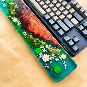 May include: A custom-made keyboard wrist rest with a resin design featuring a koi pond scene. The wrist rest has a vibrant green base with miniature koi fish, lily pads, and flowers. A mechanical keyboard with gray and black keys is next to the wrist rest.