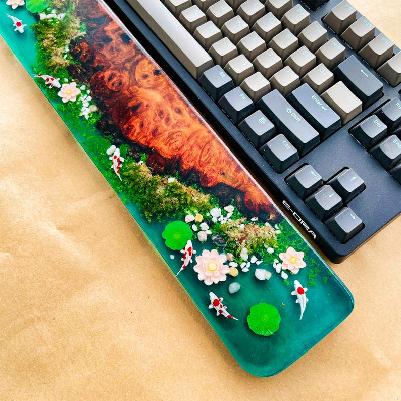 May include: A custom-made keyboard wrist rest with a resin design featuring a koi pond scene. The wrist rest has a vibrant green base with miniature koi fish, lily pads, and flowers. A mechanical keyboard with gray and black keys is next to the wrist rest.