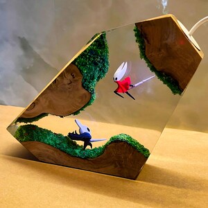 May include: A unique resin lamp featuring a diorama with miniature figures and green mossy terrain. The lamp has a wooden base and a clear resin body, showcasing the detailed figures and landscape within. The figures are in mid-flight.