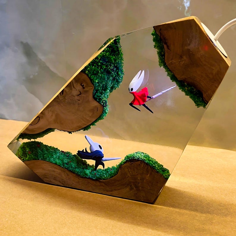 May include: A unique resin lamp featuring a diorama with miniature figures and green mossy terrain. The lamp has a wooden base and a clear resin body, showcasing the detailed figures and landscape within. The figures are in mid-flight.