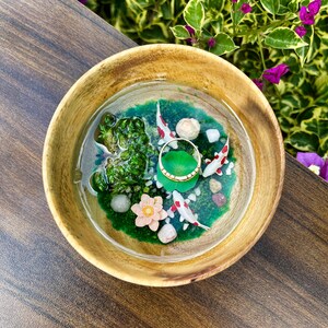 May include: A round wooden bowl containing a clear resin pond scene with green moss, small white stones, three miniature koi fish, a pink lotus flower, and a gold ring resting on a green lily pad in the center of the water.